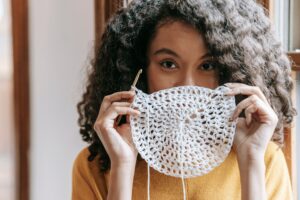 African American female with dark wavy hair wearing yellow pullover covering up mouth with white napkin with crochet and thread